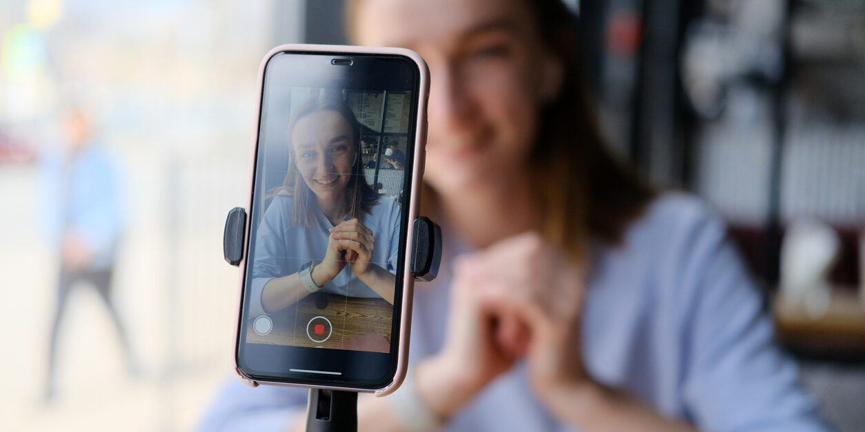 Close-up of a phone streaming video online. Blogger takes video for internet blog. A young beautiful woman blogger records a live broadcast video on her smartphone in a cozy interior cafe. Young woman record video. Online influencer girl in social media marketing live blog. Technology and online education